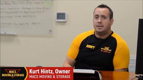 Kurt Hintz, owner of MACS Moving & Storage, sitting at an office desk, wearing a branded yellow and black shirt, discussing marketing strategies for moving services.
