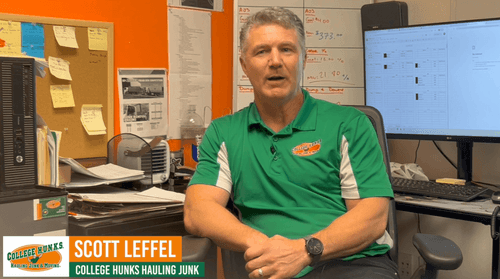 Scott Leffel speaking at his desk, wearing a College H.U.N.K.S. Hauling Junk shirt, with office supplies and documents visible in the background, emphasizing the moving and junk removal business context.