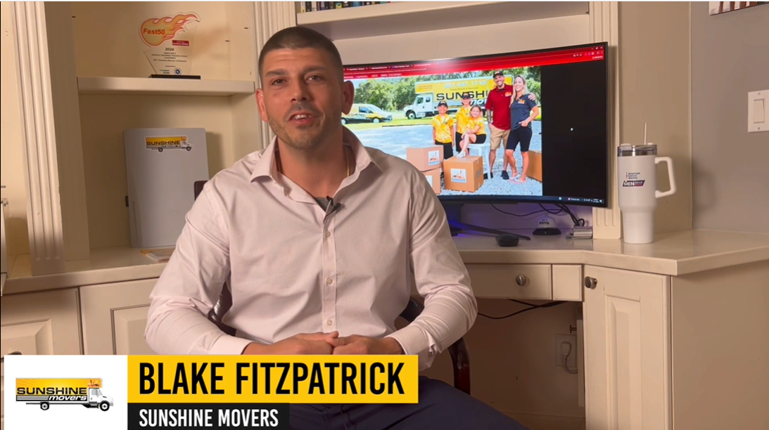 Blake Fitzpatrick sitting in an office, promoting Sunshine Movers, with a computer screen displaying the company's logo and team members in front of moving boxes.