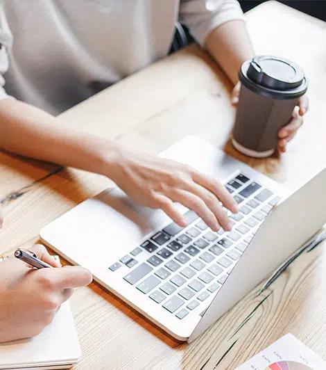 Woman holding a coffee cup and typing something on a laptop.